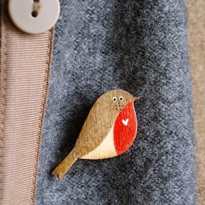 Könnte beinhalten: Ein hölzerner Rotkehl-Vogel-Anhänger mit roter Brust und einem darauf gemalten weißen Herzen. Der Anhänger ist an einem grauen Stoffhintergrund befestigt.