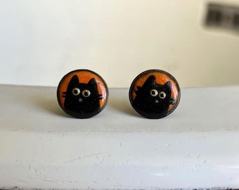 Könnte beinhalten: Ein Paar runde Ohrstecker mit einer schwarzen Katzenillustration auf leuchtend orangefarbenem Hintergrund. Das Katzendesign zeigt zwei große, runde Augen und einfache Schnurrhaare, eingefasst in einen polierten, abgerundeten Metallrahmen.