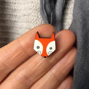 Könnte beinhalten: Ein hölzerner Fuchsanstecker mit orange-weißem Gesicht, schwarzen Augen und kleinen schwarzen Punkten. Ein kleines, dekoratives Accessoire.