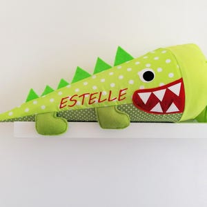 Könnte beinhalten: Ein limettengrüner, kegelförmiger Beutel mit weißen Punkten, der einem Krokodil ähnelt. Er hat grüne Filzspitzen, Füße und einen rotbezahnten Mund. Der Name "ESTELLE" ist rot gestickt. Ein lustiges, skurriles Design.