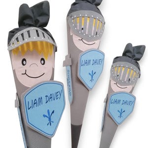 Könnte beinhalten: Drei graue und schwarze Ritter-Schultüten mit einem blauen Schild und dem Namen "Liam Davey" bestickt auf jeder. Die Schultüten sind mit einem grauen Helm und einer gelben Feder verziert.