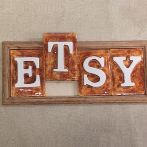 May include: A wooden frame with the word "ETSY" spelled out in white ceramic tiles. The tiles have a brown and orange glaze.