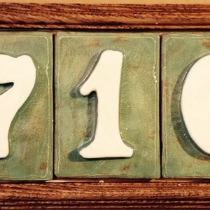 May include: A wooden frame with three white ceramic tiles displaying the numbers 716. The tiles are set against a green background.