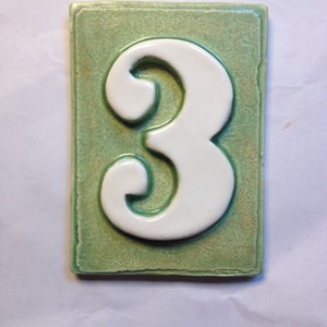 May include: A green ceramic tile with a white number 3. The tile has a textured surface.