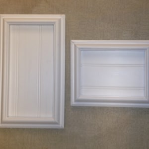 May include: Two white framed wall decor pieces with raised molding and a wood grain texture. The larger frame is vertical and the smaller frame is horizontal.
