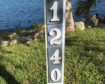 May include: White ceramic house numbers 1240 on a white post. The numbers are on a blue background. The post is in front of a green lawn and a body of water.