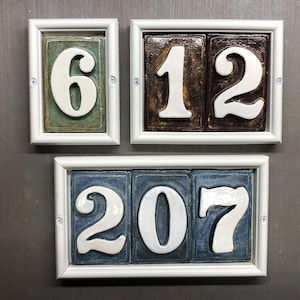 May include: Three white ceramic house numbers in white frames. The numbers are 6, 12, and 207. The frames are white with a silver trim.