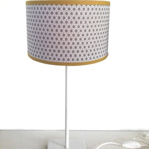 Op de afbeelding: Een witte tafellamp met een cilindrische lampenkap. De lampenkap heeft een zwart-wit geometrisch patroon en een gele rand. De lamp heeft een witte voet en een wit snoer.