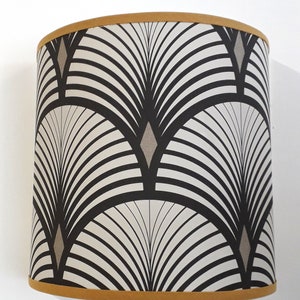 Op de afbeelding: Cilindrische lampenkap met een herhalend Art Deco-ventilatorpatroon in zwart en wit, geaccentueerd met grijze ruitvormen. De boven- en onderranden zijn afgewerkt met een goudkleurige band.