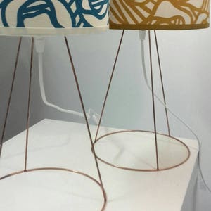 May include: Two table lamps with copper wire bases and patterned fabric lampshades. The lampshades are white with blue and yellow abstract designs.