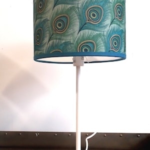 Op de afbeelding: Een witte tafellamp met een lampenkap met een patroon van pauwenveren in turquoise en groen.