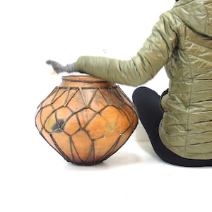 May include: A person wearing a green hooded jacket sits on the floor with their back to the camera. They are holding a large, round, woven basket with a brown and tan pattern.