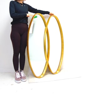 Puede incluir: Dos espejos ovalados con marcos dorados están apilados uno encima del otro. Los espejos están mirando hacia adelante y están ligeramente inclinados. Los espejos son grandes y tienen una superficie reflectante.