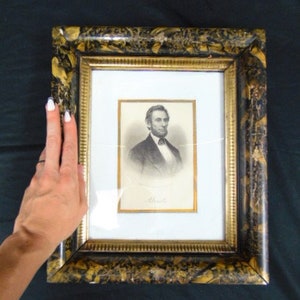 May include: A black and gold framed portrait of Abraham Lincoln. The frame has a marbled pattern with gold accents. The portrait is in black and white and shows Lincoln's face and shoulders.