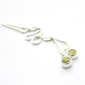 May include: A pair of silver threader earrings with two round, light green gemstones. The gemstones are set in a silver bezel. The earrings are on a white background.