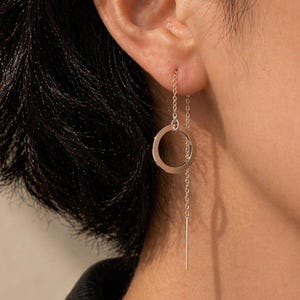 May include: A pair of delicate gold-colored threader earrings. Each earring features a small, open circle connected to a fine chain that threads through the ear. A straight bar hangs from the circle, creating a minimalist design.