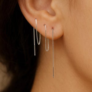 Pode incluir: São mostrados três brincos de prata, cada um com um design diferente. Um é um simples pino, outro é um pequeno aro e o terceiro é uma corrente longa que pende. Os brincos são usados na orelha.