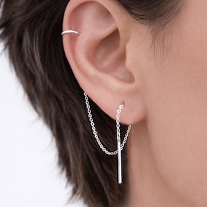 May include: A silver chain earring with a straight bar pendant. The earring is connected to the ear with a chain that drapes down to the bar. A silver hoop earring is also visible on the upper ear. The jewelry is worn on a person's ear.