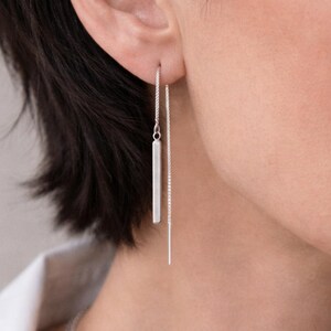 May include: A pair of silver threader earrings. One earring features a small, rectangular bar suspended from a delicate chain. The other earring has a long, straight bar. The earrings are shown against a neutral background.