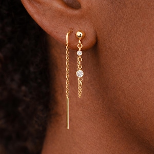 May include: A pair of gold earrings. One earring features a small gold ball and two dangling clear gemstones. The other earring has a long, thin gold bar that hangs down. The earrings are displayed on a person's ear.
