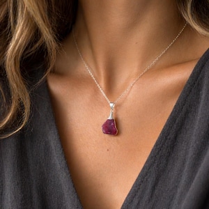 May include: A silver necklace with a pendant featuring a raw, irregularly shaped, deep red gemstone. The pendant is suspended from a delicate silver chain. The necklace is displayed against a dark background.