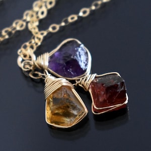May include: A gold-colored necklace featuring three gemstone pendants: a purple amethyst, a golden citrine, and a deep red garnet. Each stone is wire-wrapped and suspended from a delicate chain. The background is a dark, reflective surface.