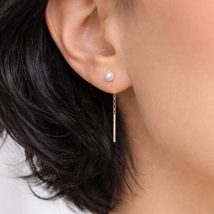 May include: A close-up of a woman's ear adorned with a delicate earring set. The earring features a small, round pearl stud and a dangling, slender silver bar. The jewelry is simple and elegant, suitable for everyday wear.