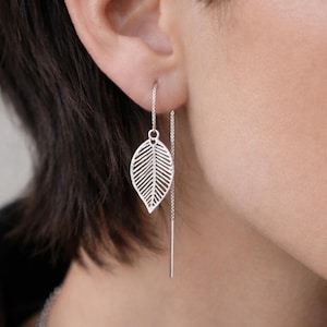 May include: Silver leaf earrings. These earrings feature a delicate, openwork leaf design suspended from a fine chain. The leaf is intricately detailed, showcasing the veins of the leaf. The earrings are a simple yet elegant accessory.