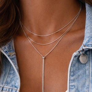 May include: A silver necklace with three delicate strands. The necklace has a sliding adjustable closure and is worn against a denim jacket. The jewelry is a simple and elegant accessory.