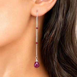 May include: Dangling silver earrings with a chain design. The earrings feature small red beads and a larger, teardrop-shaped ruby at the bottom. The earrings are shown on a person's ear.