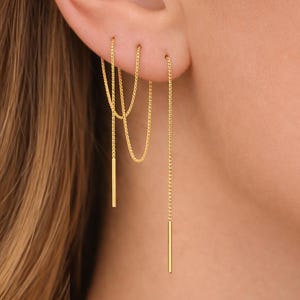 May include: Gold-colored chain earrings with a linear design. The earrings feature delicate chains that hang down, with a small bar at the end of each chain. The earrings are shown on an ear, with the chain dangling down.