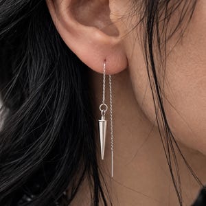 May include: A silver threader earring featuring a cone-shaped pendant and a dangling bar. The earring is shown close-up, showcasing the fine chain and the geometric design of the charm. The earring is worn in an ear piercing.