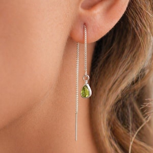 May include: A close-up of a silver threader earring with a pear-shaped, light green gemstone. The earring is dangling from the ear, showcasing the delicate chain and the vibrant color of the stone. The earring is a simple yet elegant piece of jewelry.