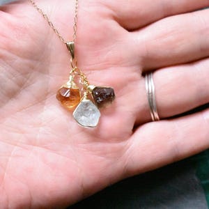 Może przedstawiać: Złoty łańcuszek z naszyjnikiem z zawieszką z trzema surowymi kamieniami szlachetnymi: jasnobrązowym, ciemnobrązowym i przezroczystym kryształem.