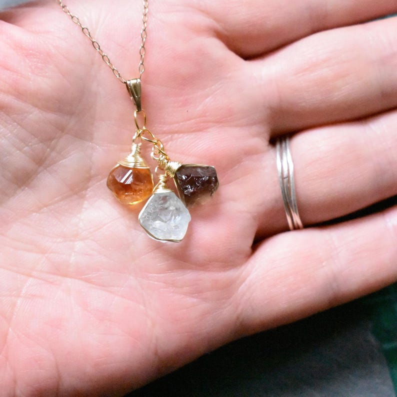 Może przedstawiać: Złoty łańcuszek z naszyjnikiem z zawieszką z trzema surowymi kamieniami szlachetnymi: jasnobrązowym, ciemnobrązowym i przezroczystym kryształem.