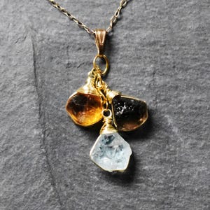 Może przedstawiać: Złoty łańcuszek z zawieszką z trzema surowymi, fasetowanymi kamieniami szlachetnymi: jasnobrązowym cytrynem, ciemnobrązowym kwarcem dymnym i przezroczystym akwamarynem. Kamienie są owinięte złotym drutem.