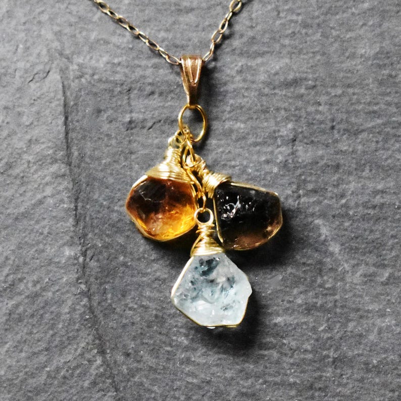 Może przedstawiać: Złoty łańcuszek z zawieszką z trzema surowymi, fasetowanymi kamieniami szlachetnymi: jasnobrązowym cytrynem, ciemnobrązowym kwarcem dymnym i przezroczystym akwamarynem. Kamienie są owinięte złotym drutem.