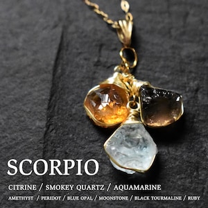 Może przedstawiać: Złoty naszyjnik z trzema surowymi kamieniami szlachetnymi: cytrynem, kwarcem dymnym i akwamarynem. Naszyjnik jest oznaczony jako "SCORPIO" i zawiera listę kamieni urodzeniowych dla każdego znaku zodiaku: ametystu, perydotu, opalu błękitnego, kamienia księżycowego, czarnego turmalinu i rubinu.