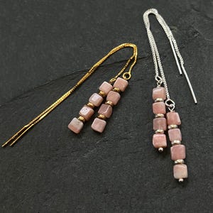 May include: Two pairs of dangle earrings. One pair features a gold chain with pink square beads and gold accents. The other pair has a silver chain with pink square beads and silver accents. The earrings are displayed on a dark gray surface.