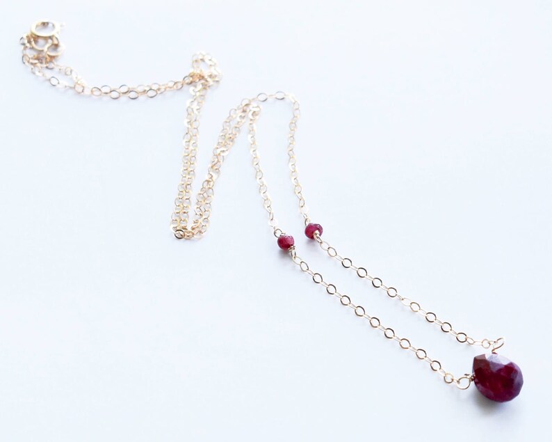 May include: A delicate gold chain necklace with a ruby pendant and two smaller ruby accents. The necklace is on a white background.