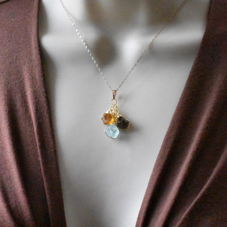 Może przedstawiać: Złoty łańcuszek z zawieszką z trzema surowymi kamieniami szlachetnymi: jasnoniebieskim akwamarynem, brązowym topazem i złotym cytrynem.