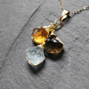 Może przedstawiać: Złoty łańcuszek z zawieszką z trzema surowymi kamieniami szlachetnymi: jasnoniebieskim akwamarynem, ciemnobrązowym kwarcem dymnym i złotym cytrynem.