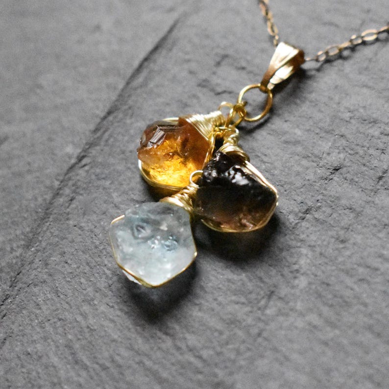 Może przedstawiać: Złoty łańcuszek z zawieszką z trzema surowymi kamieniami szlachetnymi: jasnoniebieskim akwamarynem, ciemnobrązowym kwarcem dymnym i złotym cytrynem.
