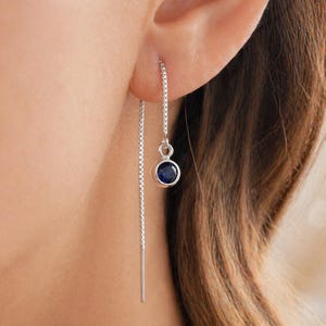 May include: A close-up of a silver threader earring with a small, round, dark blue gemstone. The earring is shown in an ear, with the chain dangling down. The gemstone is set in a silver bezel.