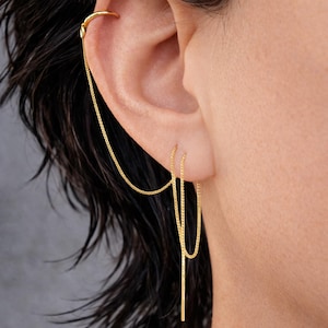 Può includere: Gioiello per l'orecchio dorato con una delicata catena che scende da un ear cuff curvo a un paio di orecchini pendenti. Il design include una barra lunga e sottile che pende sotto il lobo dell'orecchio, creando un look moderno ed elegante.