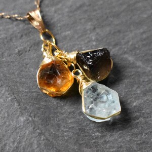 Może przedstawiać: Złoty łańcuszek z trzema surowymi zawieszkami z kamieni szlachetnych: jasnobrązowym cytrynem, ciemnobrązowym kwarcem dymnym i jasnoniebieskim akwamarynem.