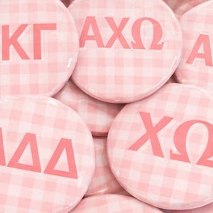 May include: A selection of round badges with a pink and white gingham pattern. Each badge displays Greek letters in pink, including ΚΚΓ, ΑΧΩ, ΔΔΔ, and ΧΩ. These are likely sorority badges.