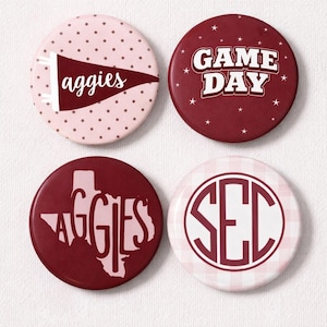 May include: Four circular badges with maroon and pink designs. One badge has the word "aggies" on a pennant, another says "GAME DAY" with stars, a third features "AGGIES" in a Texas outline, and the last has "SEC" in a circle.