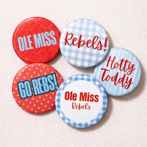 Puede incluir: Un conjunto de cinco botones redondos con texto. Los botones presentan diseños rojos, blancos y azules, incluyendo lunares, cuadros y rayas. El texto incluye "Ole Miss", "Rebels", "Go Rebs!" y "Hotty Toddy".