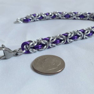 May include: A handmade bracelet featuring a Byzantine chain design. The bracelet is made of silver-colored metal links with purple accents. A silver clasp is attached. A dime is next to the bracelet.